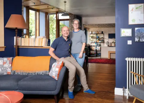 IWC Media Emily, a woman with greying hair and Robert, a hairless man, sit on a trendy denim blue sofa in their open plan living area. Bright cushions and throws in orange and white lift the mood, with an orange coffee table sitting on the polished wood floor.