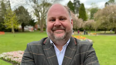 Leader of Swindon Borough Council, Jim Robbins, standing in what looks like a park. There are flower beds, tress and bushes behind him. He smiling and folding his arms in front of his chest. 