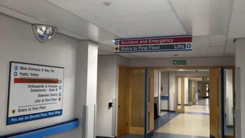 A hospital corridor which has a series of open wooden doors and a sign for Accident and Emergency hanging from the ceiling.