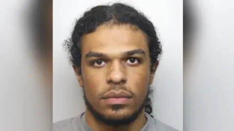 Thames Valley Police Jayden Gubetini mugshot pictured looking straight at the camera, not smiling. He has a goatee beard and hair tied back in a ponytail, brown eyes and wearing a grey sweater.