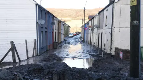 The entrance to a Cwmtillery street in daylight is blocked by rubble and mud. Beyond the rubble, water stands in the street and the mountain behind is visible in the background