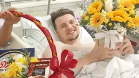 Family A man with brown hair and a small beard in a white hospital bed with a small gift with yellow flowers behind him and a hand holding a basket of food with a red bow on it.