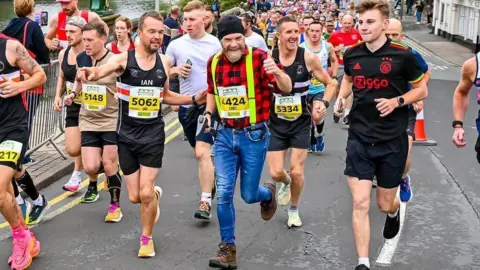 Chris Garratt A man dressed in a lumberjack outfit - a black hat, red and black shirt, yellow suspenders, jeans and brown boots - smiles and puts his thumb up as he runs past amid a crowd of runners.