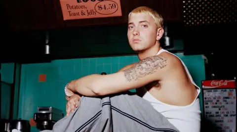 Steve Read A portrait photograph of Eminem seated indoors. He has light-coloured hair and is wearing a white sleeveless top and loose grey trousers. A tiled wall and menu board are visible in the background.