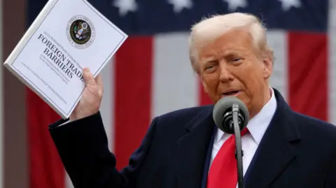 Donald Trump holds a document that says 'Foreign Trade Barriers'. He wears a blue suit, white shirt and red tie. There is a mic in front of him and a US flag behind.