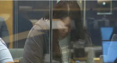 TVNZ Sullen looking woman with her head bowed as seen from behind glass