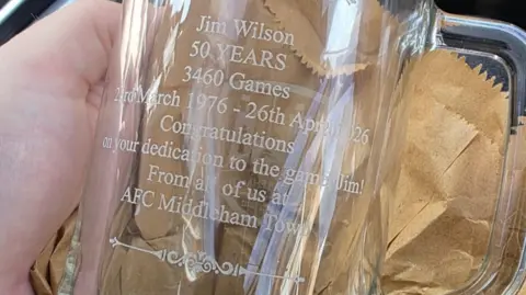 LS Photography This image is a close-up photograph of a clear glass tankard held in a hand. The engraved text appears frosted or etched. The background includes crumpled brown packing paper.