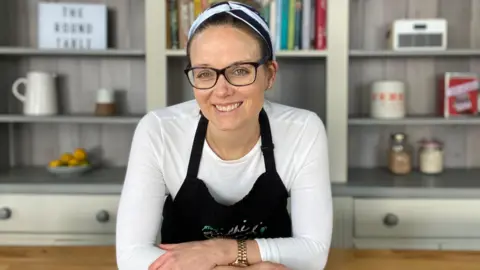 Chloe Coker wearing black-rimmed glasses, a black and white headband, a white long-sleeved top and black apron, leans forward, smiling, with her arms crossed over her wooden kitchen island. Kitchen units and shelving, with carefully arranged books and kitchenware, are in the background