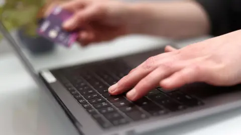 A person types on a grey laptop with a black keyboard while holding a purple debit card.