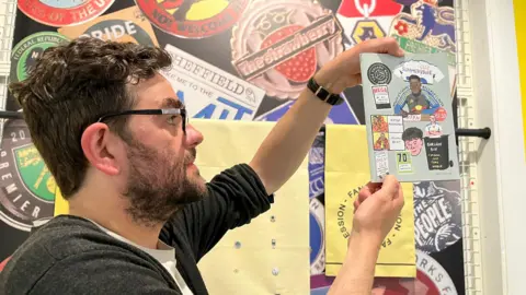 A man with dark hair, beard and glasses, wearing a grey cardigan and white T shirt holds a magazine with cartoons showing Leeds United fans