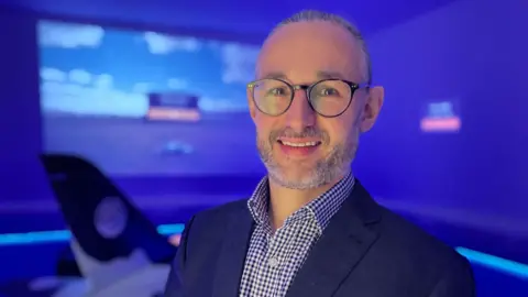 Fiona Irving/BBC Matthew Jee, Director, UK SAF Clearing House, pictured at the pilot facility in Sandwich, Kent. Matthew, who is wearing a checked shirt, suit jacket, and glasses, is smiling to camera. 
