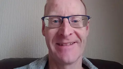 Alan Mcaulay is smiling at the camera. He is wearing glasses that have dark rims at the top. There is white wallpaper behind him