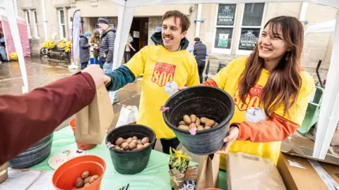 Paul Campbell A man and a woman wearing yellow T-shirts over hooded offer free seed potatoes at a street stall.
