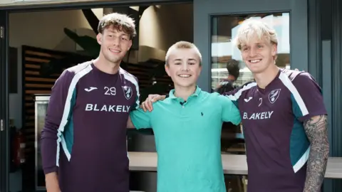 John Fairhall/BBC Ranulph, wearing a green polo shirt, is standing in between two players at the club's training ground as they pose for a photograph. Midfielders Oscar Schwartau is on the left and Pelle Mattsson is on the right, wearing Norwich City-branded maroon-coloured training tops.