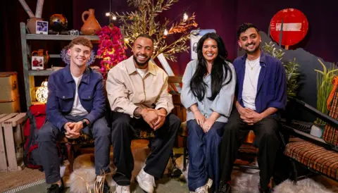 Race Across the World: The Detour Alfie Watts and Tyler West set in The Detour studio with two guests. They are all smiling at the camera and sitting in a studio with props around them such as a road stop sign, suitcases and vases. Watts is wearing a blue jacket, blue trousers and a white T-shirt. West is wearing a beige jacket, darks trousers and white trainers.  