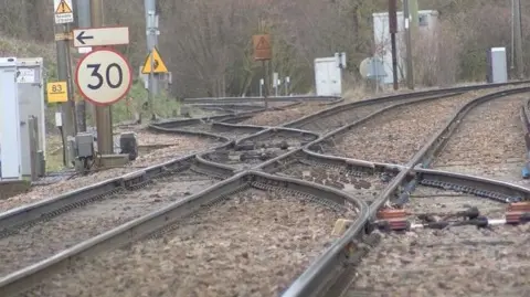 The criss-crossing railtracks at Haughley junction. The tracks of two lines curve out of view at the top of the picture. Another line is joining from the left. Various other bits of rail infrastructure are in shot, including grey track-side boxes and a sign displaying a speed limit to drivers.