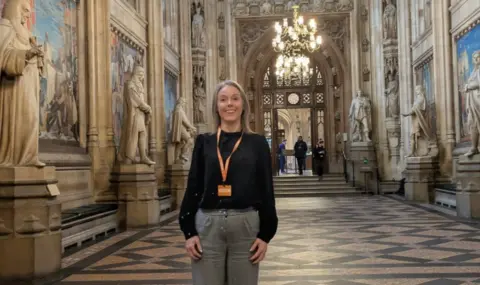 University of Wolverhampton A woman with fair hair, wearing an orange "visitor" lanyard over a black top, stands in an ornate hallway in Parliament with statues along the walls.