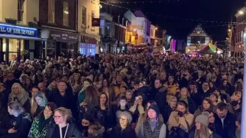 Hundreds of people stand in a street in Bridgnorth, with strings of unlit lights hanging above them. The sky is dark and there are purple lights in the background, illuminating a building