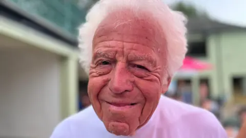 Emma Baugh/BBC Bill Marriott has short white hair. He is smiling and is wearing a white T-shirt. He is standing in front of the building.