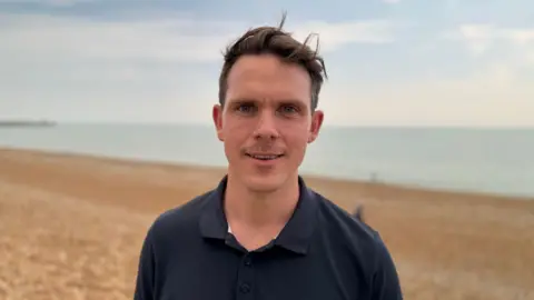 Sam Johnson who has brown short hair and a black polo top. Sam is standing on Brighton beach which has a shingle bank