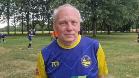 Naj Modak Michael Potts wearing a blue and yellow team shirt, standing on a grassy park field. Other players are taking part in football training drills among cones and trees in the background