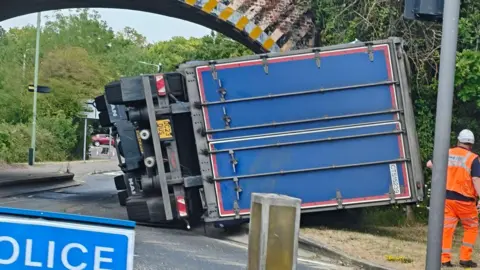 A large blue lorry that has overturned in front of a railway bridge. Surrounding cars are stationary.