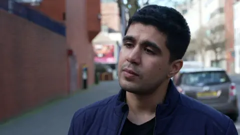 Mohammad has short black hair. He's wearing a navy coat and a black top. Cars are parked behind him. He's standing on a street. 