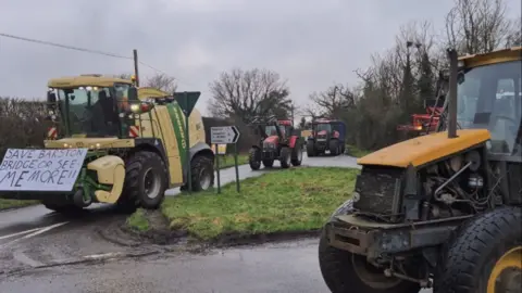 Four tractors. There are two yellow tractors, one of them holds a sign that says "save Barston bridge or see me more". There are two red tractors in the distance