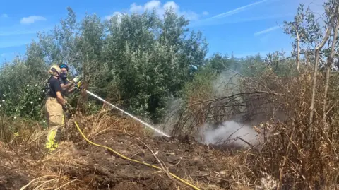 Photograph of a firefighter tackling a blaze at an illegal waste sump in Ashton-upon-Mersey. The image shows a jet of water being fired at the ground. 