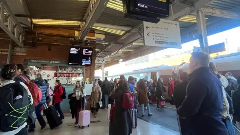 BBC/Grace Parnell People mill around and wait on a train platform at Doncaster Station. Many of them have bags and suitcases.