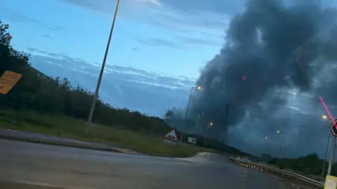 Wide view of a large black cloud of smoke rising in the air behind a road. The sky is dark as the sun begins to rise.