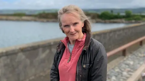 A woman with greying blonde hair in a dark jacket with a pink fleece underneath stands in front of Stranraer harbour