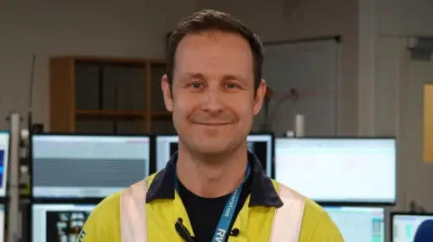 Shaun Whitmore/BBC Sam Woodgate, a man who is standing in front of a range of lit computer monitors. He is looking directly at the camera and smiling. He is wearing a fluorescent jacket and RWE blue lanyard round his neck. 