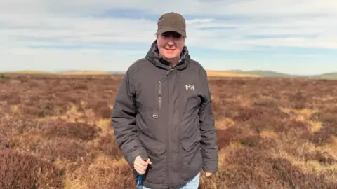 Prof Swindles is standing on a peat bog. He is smiling at the camera and is wearing a brown cap. He has a brown waterproof coat on with HH branding on the right lapel. He is holding a walking pole.