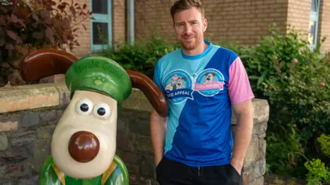 The Grand Appeal Nick Flynn is standing next to a Gromit sculpture outside the hospital in Bristol. He is smiling at the camera.