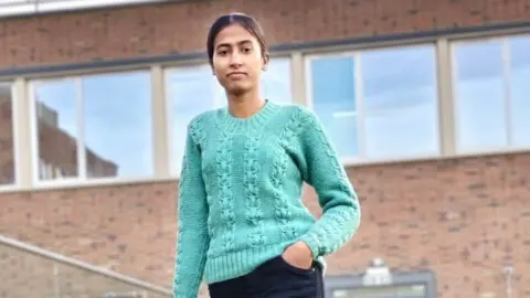 A woman in a teal knitted jumper and dark trousers stands outside in front of a red brick building. She has brown hair that has been tied back.