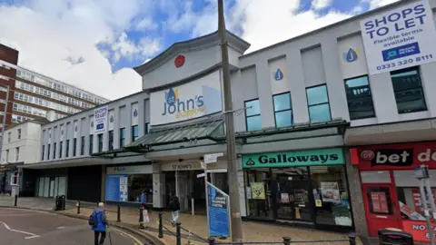 Google Street view of St John's Shopping Centre, a tired grey rendered two-storey building with shabby shops on the ground floor.