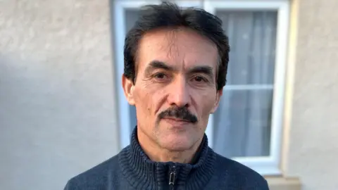 A head and shoulders image of a man with short black hair, a moustache and a dark blue zipped-up fleece. He is looking at the camera. Behind him is a window with a net curtain.