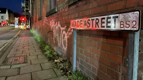 A view of Wade Street. with a street sign in the foreground and roadworks in the distance, there is graffiti on the redbrick wall.
