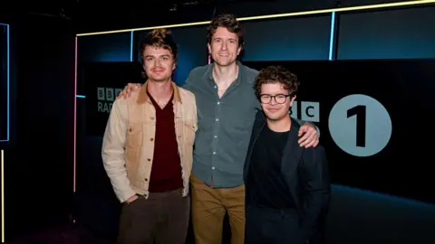 Joe Keery and Gaten Matarazzo of Stranger Things stand either side of Greg James in the BBC Radio 1 studios.