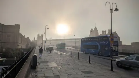 BBC Weather Watcher/Patsee A full sun is rising through mist above Bristol's Temple Meads station. The picture is semi-dark and gloomy. There is a blue double-decker bus to the right and a green single deck bus behind. Two solitary pedestrians are walking up the approach road towards the station entrance in the distance 