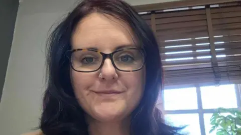 Sophie Carpenter A woman with dark purple hair and glasses sitting in front of a window.