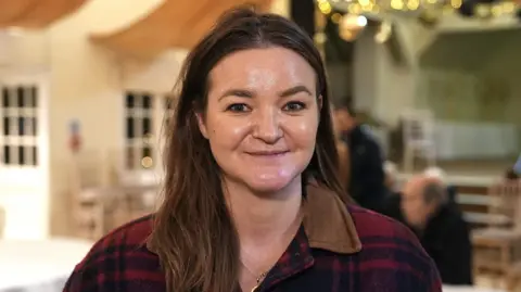 Martin Giles/BBC Jenny Riddell-Carpenter smiles at the camera. She has long brown hair and wears a red and black chequered coat.