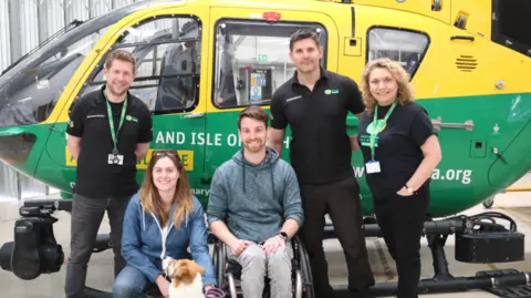Hampshire and Isle of Wight Air Ambulance Dale is sitting in a wheelchair and smiling at the camera. He is wearing a blue hoodie and grey trousers. His wife Rachael is kneeing down next to him and is wearing a blue jumper and jeans. The Hampshire and Isle of Wight Air Ambulance team of three are all dressed in black.
