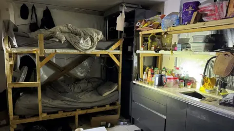 Cambridgeshire Police Bunk beds are in the corner of a kitchen area inside a house. They are scruffily-built and have untidy bedding on them. There are dark-coloured kitchen cabinets and a sink next to them. The worktops and shelves are stacked with food and plastic containers