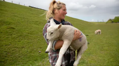 Hollie is wearing a red and black and white checker shirt and holding a lamb 