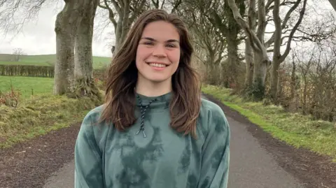 Morgan is smiling at the camera. She has long brown hair and is wearing a green hoodie. The trees can be seen behind her.