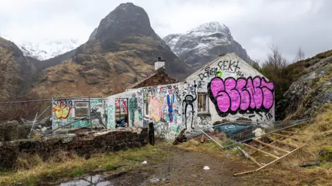 The cottage is a ruin and its white walls are covered in graffiti. Behind the ruins loom large high mountains. 