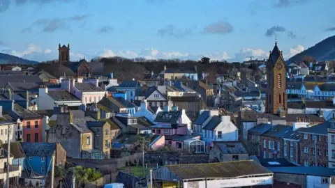 MANX SCENES A high up view of rooftops with two church towers and a hill in the background with a clear blue sky.
