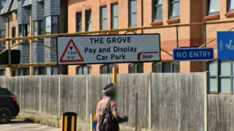 Sign for the entrance to the Grove Car Park in Slough showing height restrictions. Someone is walking past and their face is blurred out.